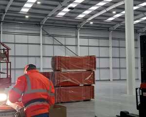 A worker in a fluorescent-lit warehouse is assembling equipment near stacked materials.