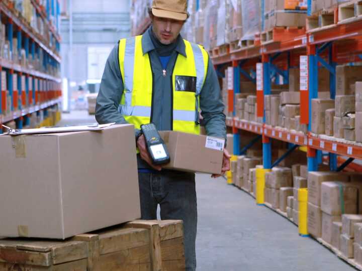Warehouse worker scanning boxes with a handheld device among shelves of packaged goods.