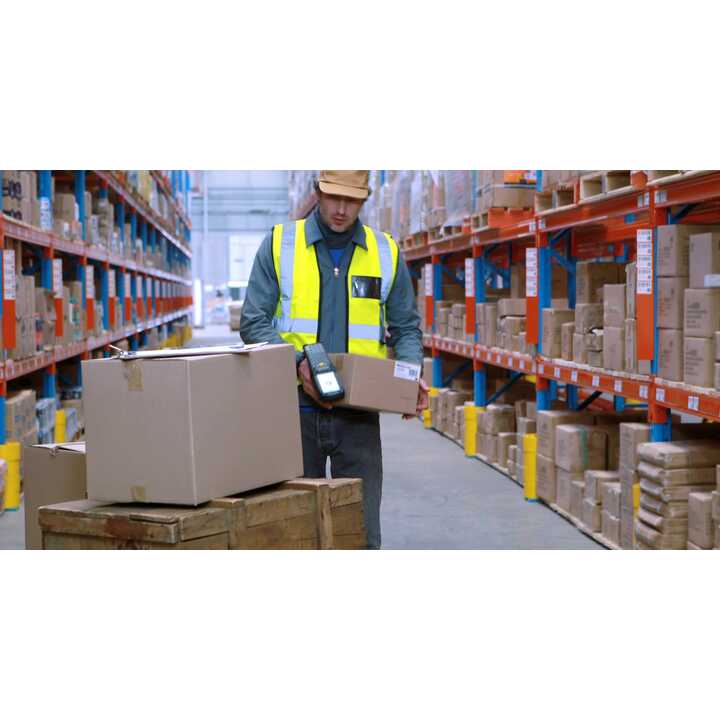 A worker in a yellow vest scans boxes in a warehouse aisle filled with shelves.