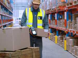 Warehouse worker scanning boxes with a handheld device among shelves of packaged goods.