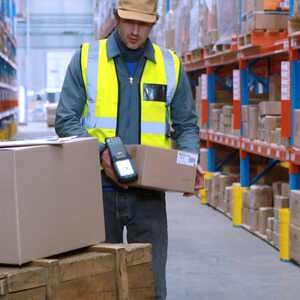 A worker in a yellow vest scans boxes in a warehouse aisle filled with shelves.