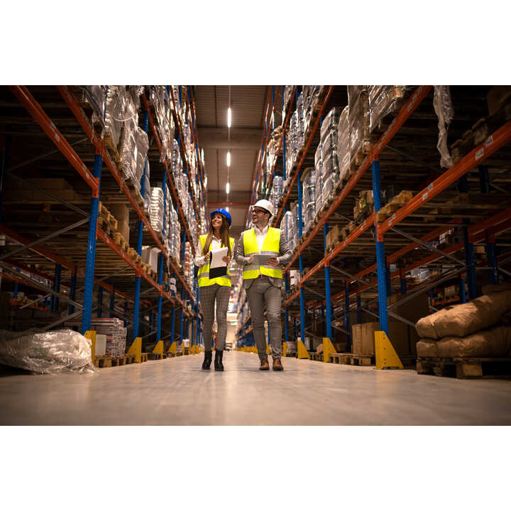 Two workers in safety vests and helmets walk through a warehouse aisle with stacked goods.