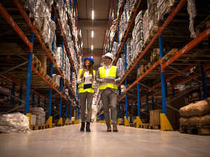 Two workers in safety vests and helmets walking through a warehouse aisle with stacked goods.