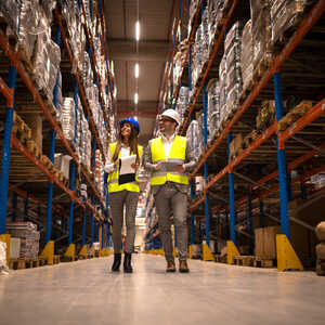 Two workers in safety vests and helmets walk through a warehouse aisle with stacked goods.