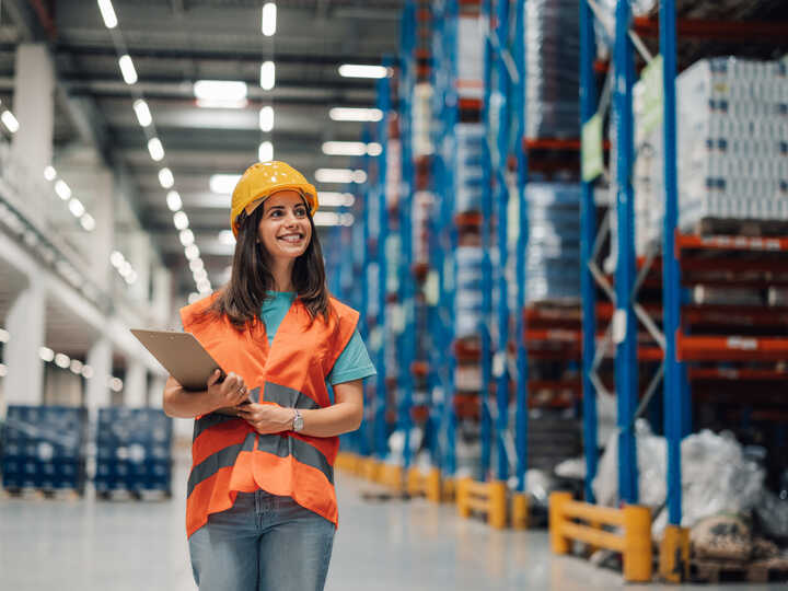 A smiling woman in a safety vest and hard hat stands in a warehouse aisle.