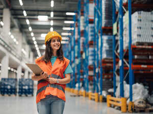 A smiling woman in a safety vest and hard hat stands in a warehouse aisle.