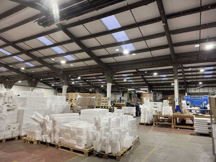 Two workers in safety vests and helmets walking through a warehouse aisle with stacked goods.