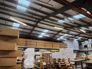 Before: Outdated lighting system in a Wiltshire factory Interior of a warehouse with stacked boxes and large windows overhead.