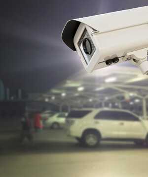 Security camera overlooking a dimly lit parking lot with parked cars and people.